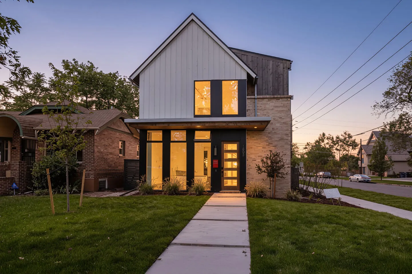 Modern two-story farmhouse at dusk with lit floor-to-ceiling windows, brick accents, and a front walkway